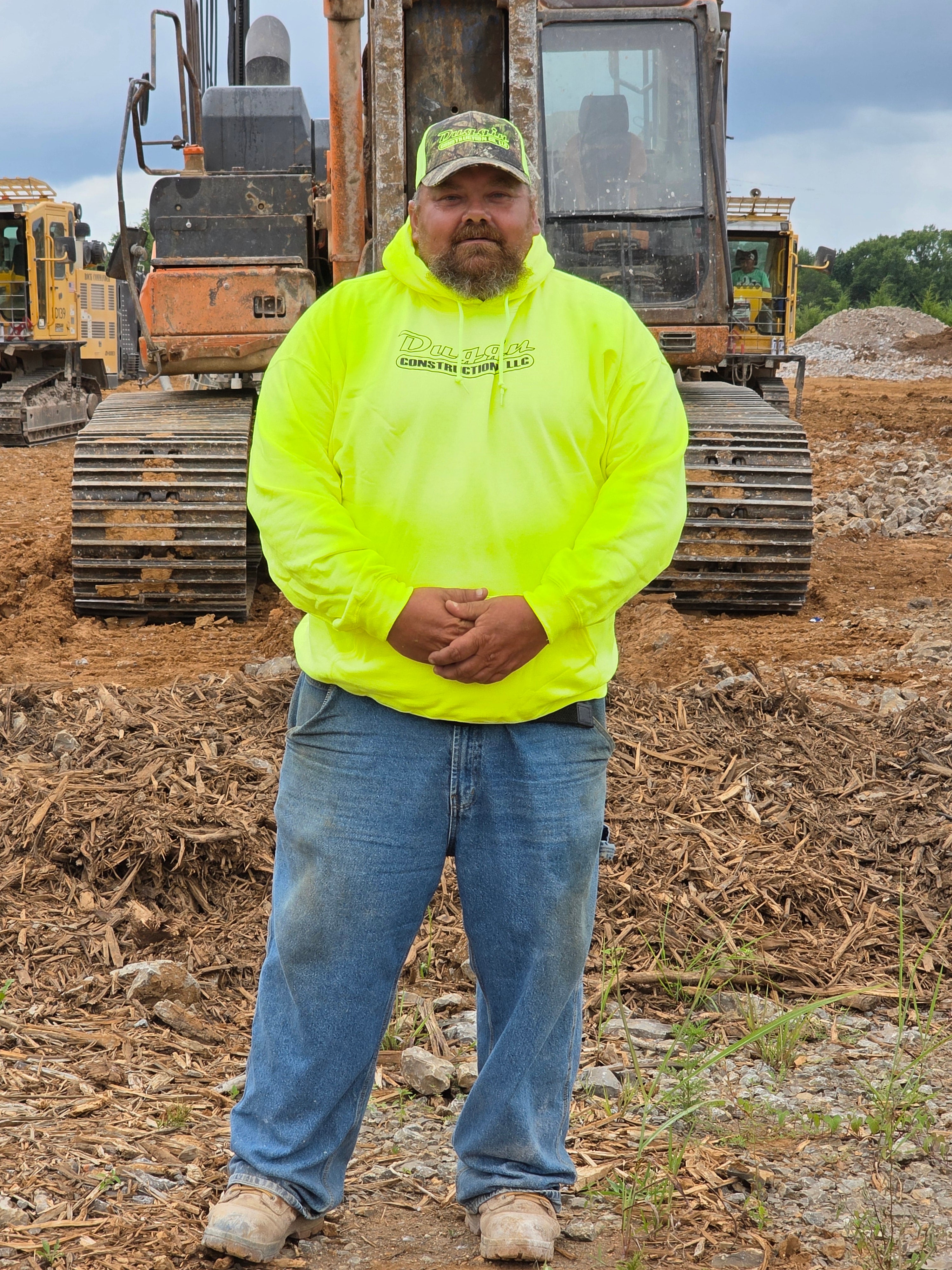 Person wearing a bright yellow safety jacket and blue jeans standing on a construction site with machinery in the background.