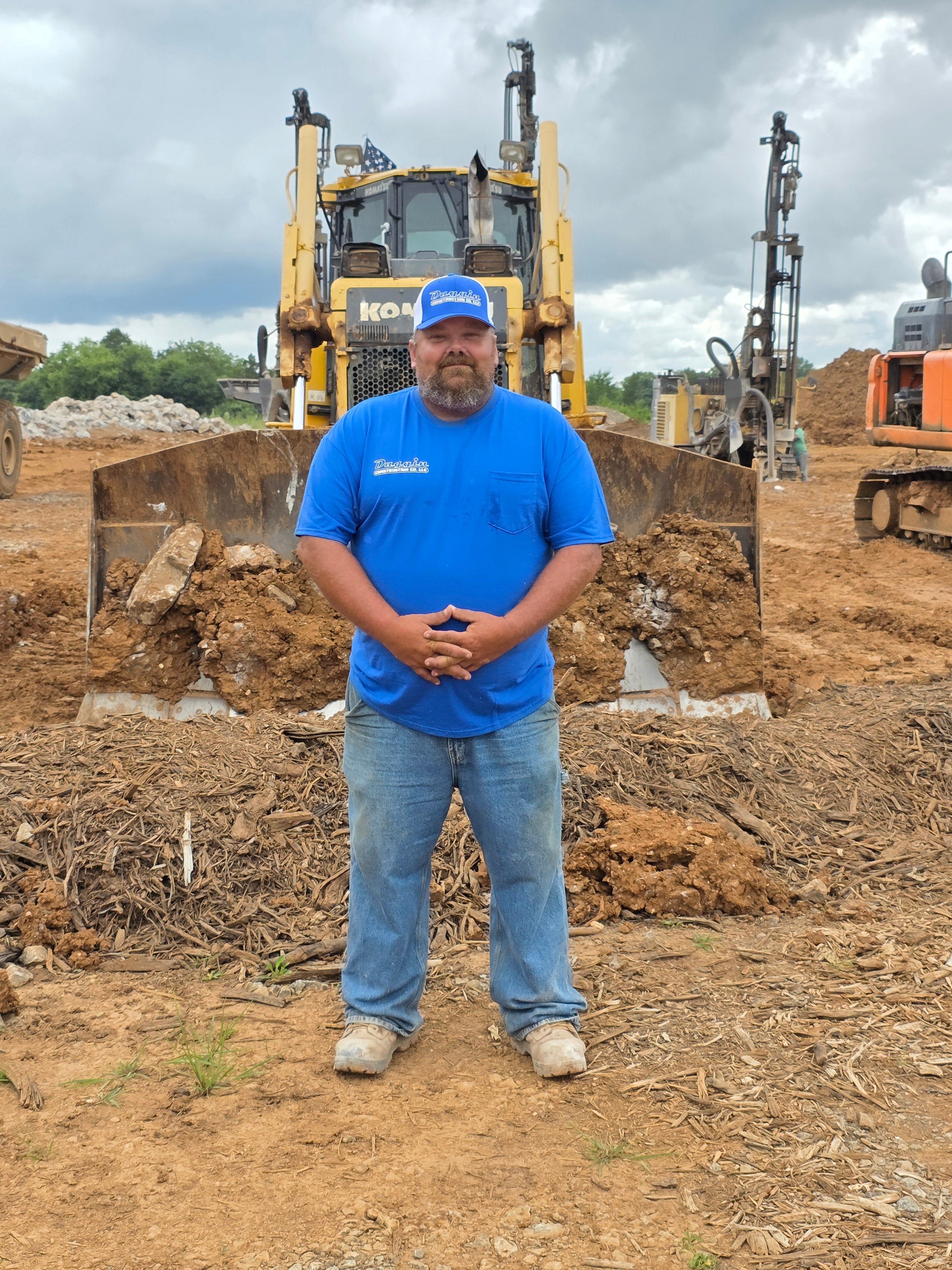 Man standing in front of dozer in a blue short sleeve shirt with and blue and white cap. 